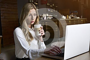 Beautiful happy woman working on laptop computer during coffee break in cafe bar. Young blonde girl in white blouse