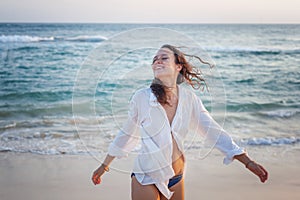 Beautiful happy young brunette woman in a white shirt enjoys enjoying life near the ocean at sunset