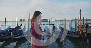 Beautiful happy young Caucasian travel blogger woman walks along empty sunrise Venice gondola pier looking at camera.