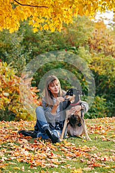 Beautiful happy young Caucasian woman sitting on ground among autumn fall yellow leaves and hugging her pet dog