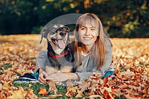 Beautiful happy young Caucasian woman sitting on ground among autumn fall yellow leaves and hugging her pet dog