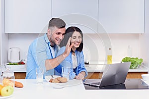 Beautiful happy young couple having breakfast at home and uses laptop at kitchen. Enjoying free time together.