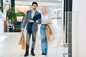 Beautiful happy young couple holding shopping paper bags with purchases and walking holding hands at mall.