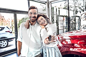 Beautiful happy young couple hugging holding the keys to their new car smiling joyfully at the dealership