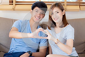 Beautiful happy young couple sitting on sofa fun making gesture heart shape with hand indoor together