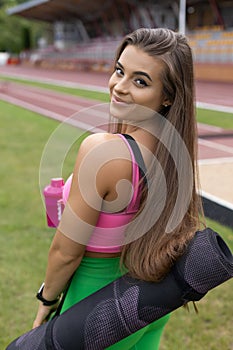 Beautiful happy young female athlete in sports uniform smiles , holds yoga mat, bottle of water.