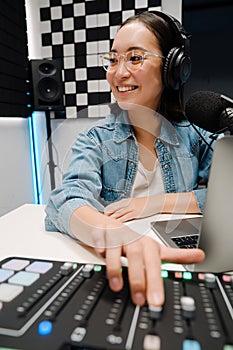 Happy young female radio host using headphones and sound mixer while broadcasting in studio