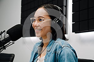 Happy young female radio host using microphone and headphones while broadcasting in studio