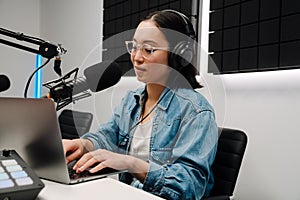 Beautiful young female radio host using microphone and laptop while broadcasting in studio