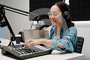 Happy young female radio host using microphone and sound mixer while broadcasting in studio