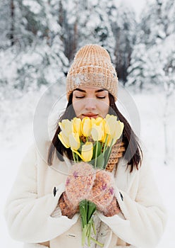 A beautiful happy young girl in gloves and a winter hat-scarf holds a bouquet of yellow tulips. Winter forest landscape