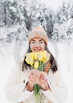 A beautiful happy young girl in gloves and a winter hat-scarf holds a bouquet of yellow tulips. Winter forest landscape