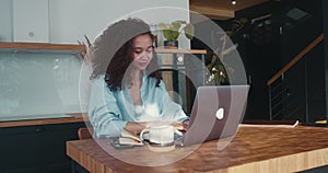 Beautiful happy young multiethnic entrepreneur business woman with curly hair taking notes, using laptop at home table.