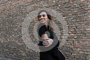 Beautiful happy young woman in an attractive smile in a stylish black t-shirt in a trendy coat posing near a brick vintage wall