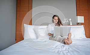 Beautiful happy young woman on bed in hotel room working on laptop, vacation business trip remote work digital nomad concept