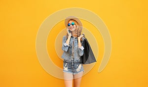 Beautiful happy young woman calling on phone with shopping bag looking away wearing summer straw hat, denim jacket on yellow