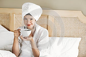 Beautiful happy young woman drinking coffee or tea in bed in hotel room or home bedroom. Fashionable  brunette girl in white towel