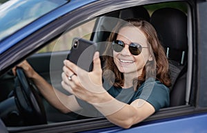 Beautiful happy young woman driver sitting in car wearing sunglasses using smartphone