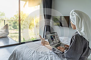 Beautiful happy young woman in a gray bathrobe and with a towel on her head on a bed in a hotel room working on a laptop, vacation