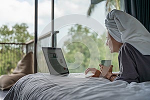 Beautiful happy young woman in a gray bathrobe and with a towel on her head on a bed in a hotel room working on a laptop, vacation