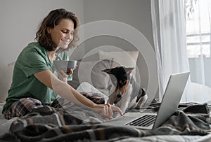 Beautiful happy young woman at home having breakfast on bed in pajamas in front of laptop with african terrier bassenji dog