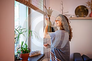 Beautiful happy young woman opening rolled bamboo blinds in country house in summer morning