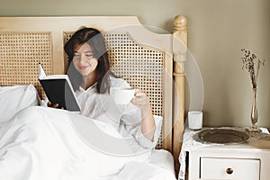 Beautiful happy young woman reading book and drinking coffee in bed in hotel room or home bedroom. Stylish brunette girl in white