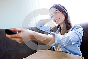 Beautiful happy young woman watching TV and holding remote control at home.
