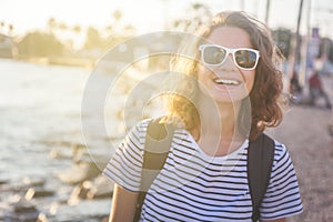 Beautiful happy young woman with wavy hair in sunglasses is laughing for joy near the sea in the rays of the sun