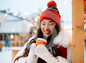 Beautiful happy young woman in winter clothes at christmas market drinking coffee