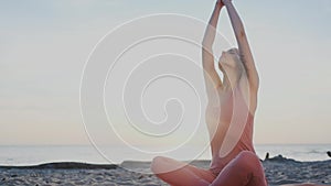 Beautiful and healthy female sits in pose of lotus on beach at sunset, practicing yoga, rise hands to the sky, breathe