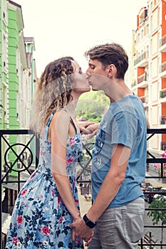 Beautiful heterosexual couple kisses on the balcony. Multicolored urban houses on the background.