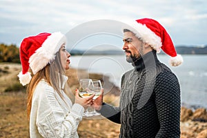 Beautiful heterosexual couple in love toasting outdoor wearing Santa Claus hat looking into each other`s eyes holding white wine