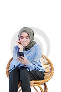 Beautiful Hijab woman sitting in a chair while holding a phone with smiling expression isolated on white backgroundan