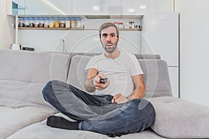 Beautiful hilarious young man holding a remote control. During this, the TV is watching while sitting on the couch at