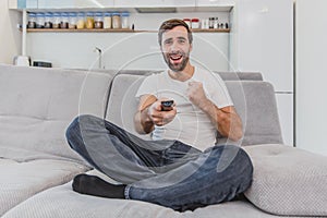 Beautiful hilarious young man holding a remote control. During this, the TV is watching while sitting on the couch at