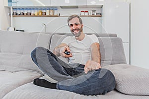 Beautiful hilarious young man holding a remote control. During this, the TV is watching while sitting on the couch at