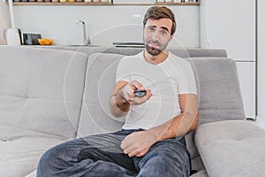 Beautiful hilarious young man holding a remote control. During this, the TV is watching while sitting on the couch at