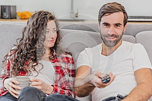 Beautiful hilarious young man holds a remote control, wife`s cup of coffee. During this man looks at the camera and