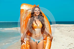 Beautiful hippie girl portrait by orange Inflatable pool float mattress on tropical beach. Fit Brunette with long healthy curly