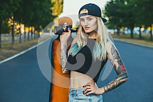 A beautiful, hipster blonde with blue hair in tattoo stands with a longboard