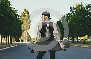 beautiful, hipster blonde with blue hair in tattoo stands with a longboard it is turned back on the street.