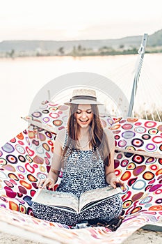 Beautiful hipster girl with big book relaxing in hammock on the beach in summer evening light. happy woman in straw hat resting