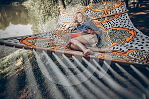 Beautiful hipster girl lying on a hammock and reading a book, camping leisure, stylish woman resting outdoors near a lake on a