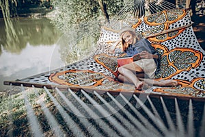Beautiful hipster girl lying on a hammock and reading a book, camping leisure, stylish woman resting outdoors near a lake on a