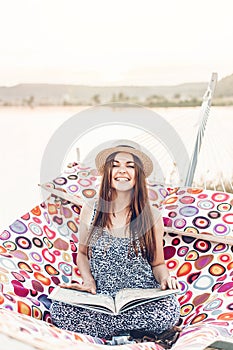 Beautiful hipster girl reading book and relaxing in hammock on the beach in summer evening light. happy woman in straw hat resting