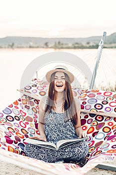 Beautiful hipster woman relaxing in a hammock, emotional girl in stylish hat reading book while resting on the beach at sunset