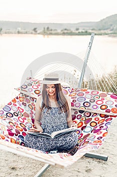 Beautiful hipster woman relaxing in a hammock, emotional girl in stylish hat reading book while resting on the beach at sunset