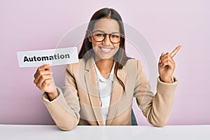 Beautiful hispanic business woman holding automation word on paper smiling happy pointing with hand and finger to the side