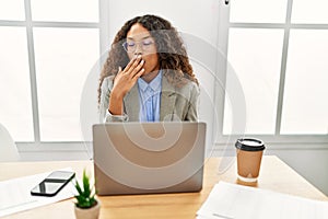 Beautiful hispanic business woman sitting on desk at office working with laptop bored yawning tired covering mouth with hand